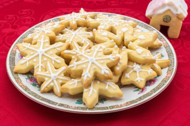 Kar tanesi şeklinde şekerli Noel kurabiyesi tabağı. Dekorasyon olarak ev şeklindeki kurabiyeyle kırmızı bir masa örtüsüne atılmış..