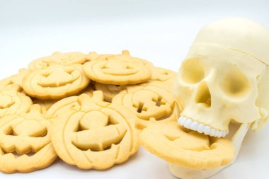 Kafatası beyaz bir arka planda, bir yığın kurabiyenin yanında balkabağı şeklinde bir kurabiyeyi ısırıyor..