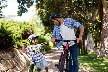 Baba oğul parkta bisikletle dikiliyorlar.