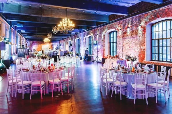 Celebration hall with tables set for banquet in loft. Vintage room with brick walls without plaster or wallpapers. Table decorated with candles, fabric and flowers.