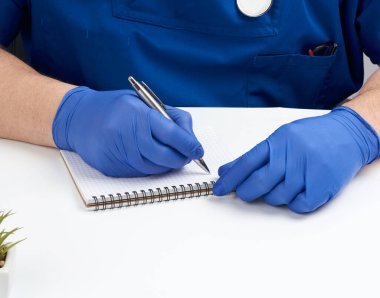 Mavi üniformalı erkek doktor beyaz bir masaya oturur ve şöyle yazar: 