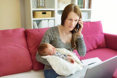 Young mom works remotely from home office with newborn baby. Freelancer woman holding her infant while using laptop. Workplace in living room. Working during quarantine