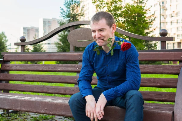 A man with a rose in his teeth sits on a park bench and looks at women