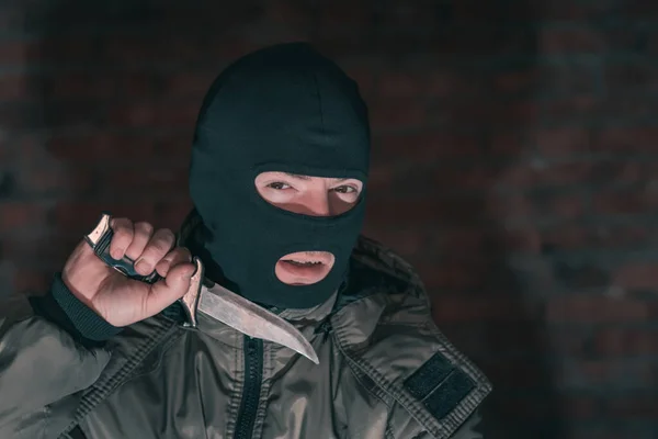A thug in a balaclava with a knife against a brick wall in the dark shows threatening gestures