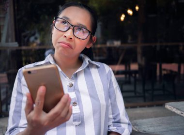 Asyalı kadınlar ahşap sandalyede akıllı telefonlarıyla otururlar ve mutsuz üzgün yüzleriyle umursamazlar, kahve kafesinde yaşam tarzlarıyla poz verirler..