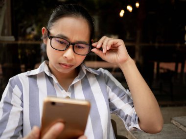 Stresli Asyalı kadınlar ahşap sandalyede akıllı telefonlarıyla oturuyorlar. Akıllı telefona uzun süre baktığı için göz ağrısı çekiyor. Sağlık ve Tıp Konsepti