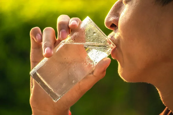 Asian teen boy drinking cool water in clear glass over blurred nature background