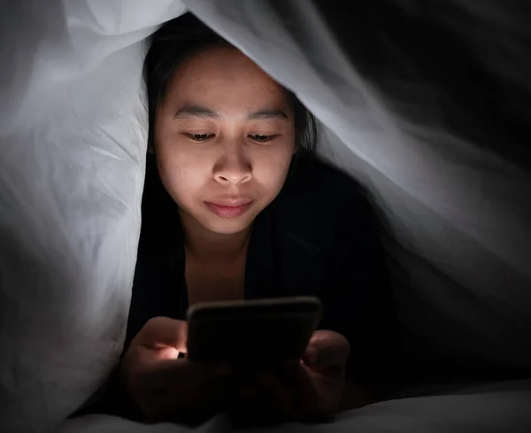 スマートフォンを使用したアジア女性の肖像画は ベッドの上の白い毛布の下に横たわっています 彼女の顔は明るいモニターで照らされている くつろぎの時間 — ストック写真