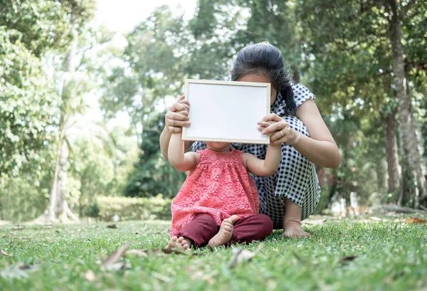 Asian little girl holding empty white board with mom and sitting on grasses ground. Space for write your text