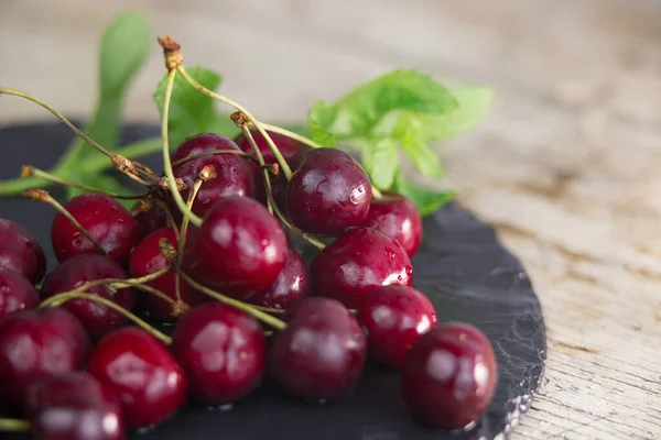Fresh cherries on a black plate of wet slate with sprig of fresh green mint on light wooden table in selective focus for copy space