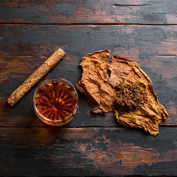 stock image Cut tobacco and tobacco leaves with cigar and whiskey rum on wood background on vintage dark table. overhead shot top view