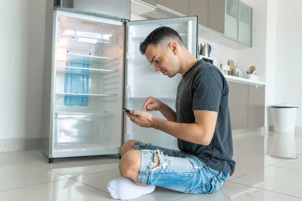 若い男がスマートフォンを使って食べ物を注文する 食べ物のない空の冷蔵庫 フードデリバリーサービス広告 — ストック写真