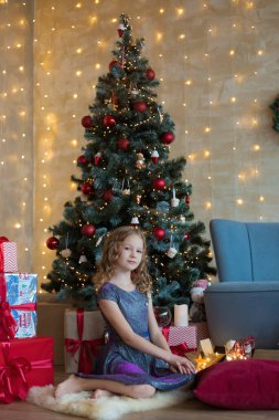 Pretty little child is sitting in front of christmas tree among garlands