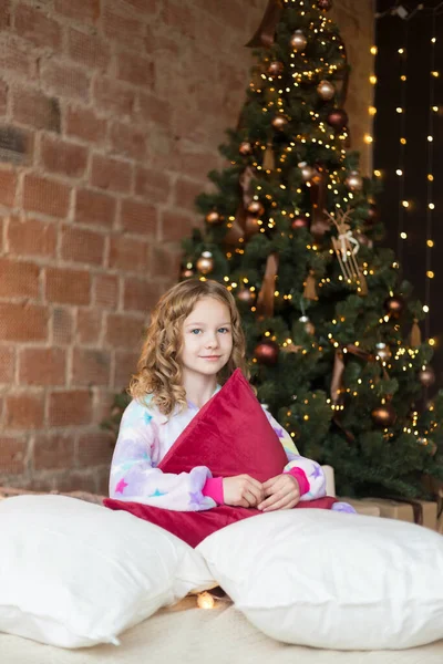 Cute little girl sits on the bed and hugs pillow on Christmas background