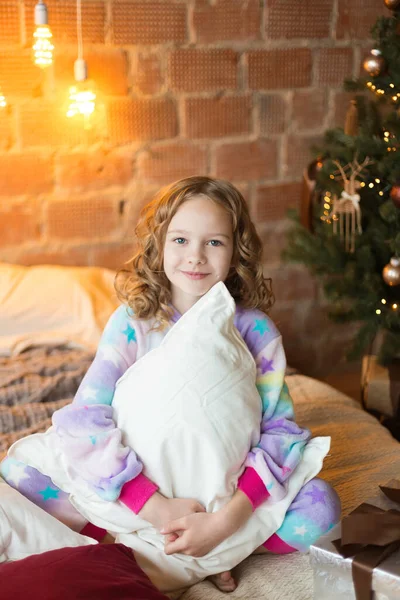 Cute little girl sits on the bed and huggs pillows with background of tree