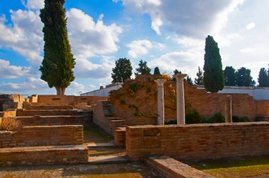 Kahverengi tuğla duvarlar, beyaz sütunlar ve yeşil selvi ağaçları. Mavi gökyüzü ve arka plan olarak beyaz bulutlar