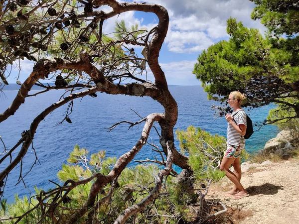 クロアチアの海辺で安心して一人で泳ぐリモートコーブを探している松の木の間の海岸沿いのパスを歩いて小さなバックパックを身に着けている若い活発な女性観光客 旅と冒険のコンセプト — ストック写真