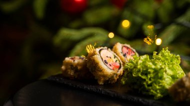Nori, taze somon, ton balığı, avokado, masago havyarı, ananas sosuyla süslenmiş salata, Noel için ahşap bir masada siyah bir tabağa süsleme olarak serpilmiş özel suşi rulosu.