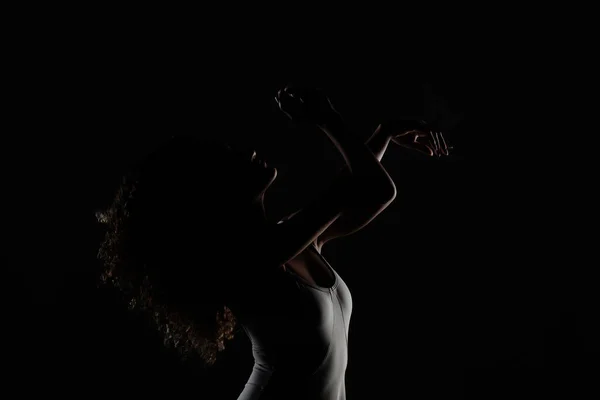 巻き毛のバレエポーズの女の子 サイドライトシルエットのバレリーナで白いドレスに黒の背景 — ストック写真