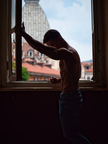 紧靠在窗边 穿着牛仔裤 向外张望的性感无袖肌肉年轻人 — 图库照片