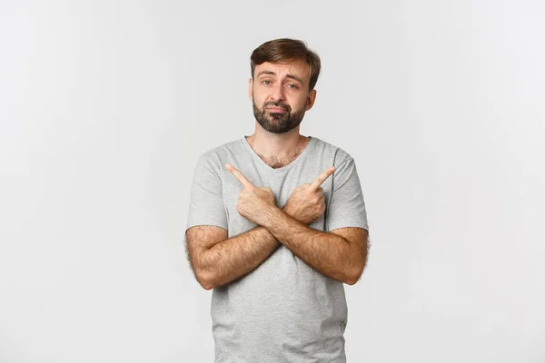 stock image Portrait of sad and confused bearded guy in gray casual t-shirt, pointing fingers sideways, showing left and right promo, standing over white background