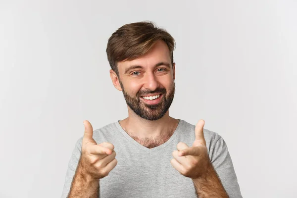 大胡子英俊男子的特写镜头 微笑着 用手指指着相机 穿着灰色T恤站在白色背景上 — 图库照片