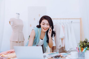 Güzel bir terzi atölyesinde çalışırken dizüstü bilgisayar ve telefon kullanıyor. Dizüstü bilgisayar kullanan terzi.