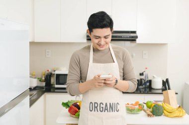 Yakışıklı Asyalı adam mutfakta akıllı telefonuyla mesaj atıyor.