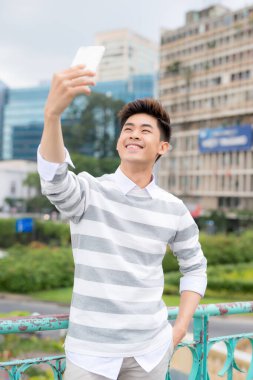 Neşeli Asyalı adam modern akıllı telefondan selfie çekerken halka açık parkta keyifli yürüyüşler yapıyor.