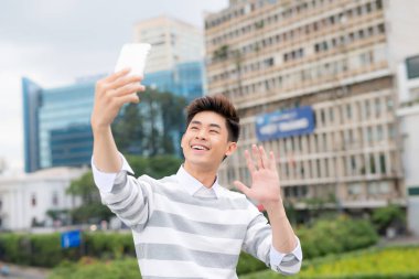 Neşeli Asyalı adam modern akıllı telefondan selfie çekerken halka açık parkta keyifli yürüyüşler yapıyor.