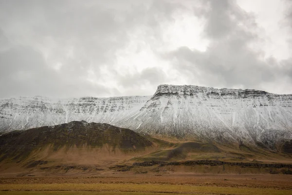 Heavenly light through the clouds over a snow capped mountain ridge leading down into brown grassy landscape in Iceland