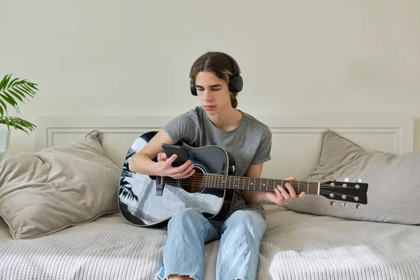 Adolescente Masculino Fones Ouvido Sentado Casa Com Guitarra Acústica Smartphone — Fotografia de Stock