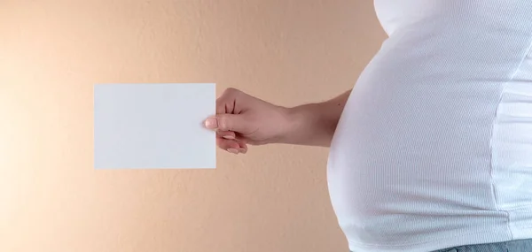 A close-up view of the belly of a pregnant woman in a white T-shirt that is holding an empty sheet of paper.