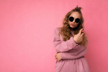 Güzel, mutlu, esmer, kıvırcık saçlı bir kadının pembe arka plan duvarından izole edilmiş, günlük pembe spor kıyafetleri ve şık güneş gözlükleriyle kameraya bakıp eğlendiği bir fotoğraf.