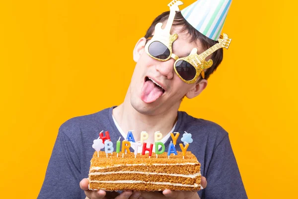キャップと燃えるキャンドルと黄色の背景にポーズをとる彼の手の中に自家製ケーキを持つ肯定的な面白い若いアジアの男 記念日と誕生日のコンセプト — ストック写真