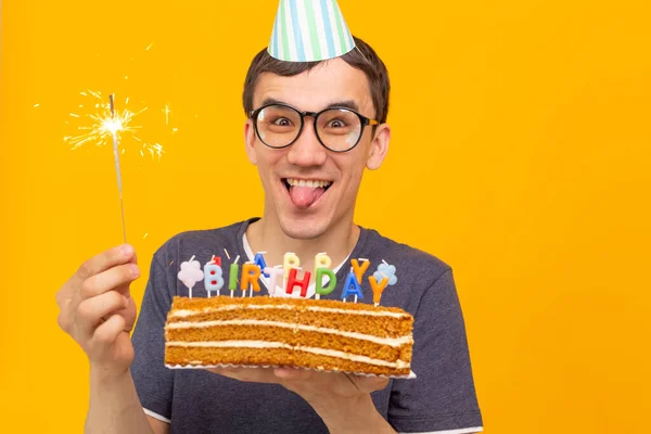 彼の手に燃えるろうそくと黄色の背景に祝福の自家製ケーキを持つメガネを持つクレイジー陽気な若いアジアの男 誕生日と記念日のお祝いのコンセプト — ストック写真