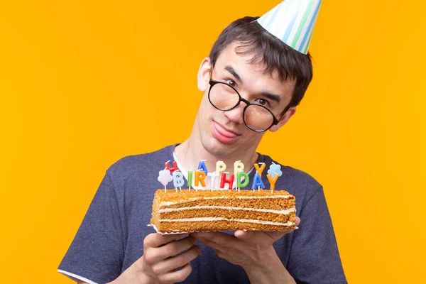 黄色の背景に彼の手の中でお祝いの自家製ケーキを保持している紙のキャップとメガネを持つ面白い肯定的な男の肖像画 コンセプトと楽しさとお祝い — ストック写真
