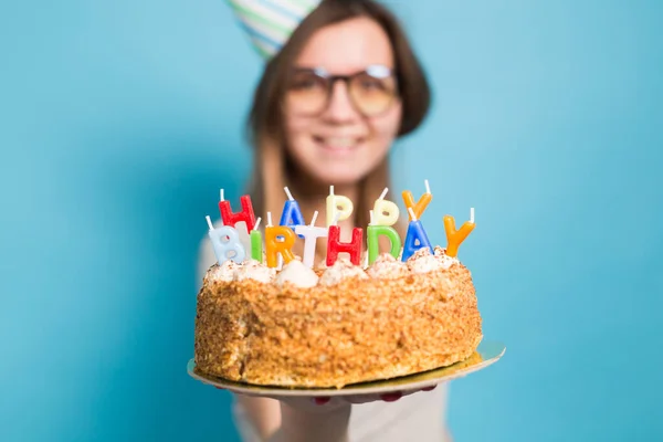 青い背景に立って彼女の手に幸せな誕生日ケーキを保持し お祝いの紙の帽子で魅力的な陽気な狂気の若い女の子の学生 広告スペース — ストック写真