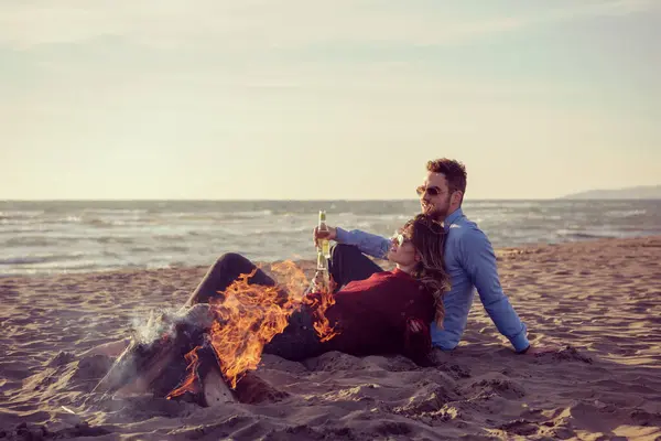 坐在海滩边篝火边喝啤酒的年轻夫妇 — 图库照片