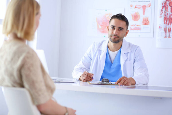 Красивый врач разговаривает с молодой пациенткой и делает заметки, сидя в своем кабинете.