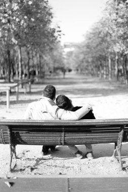 Black and white photo, back view of two hugging gays sitting on bench in park clipart