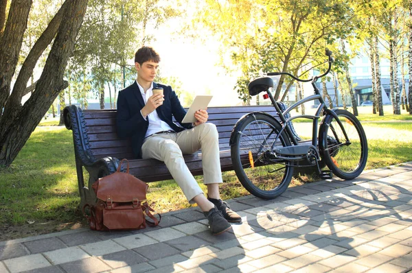 商人在喝咖啡休息 他坐在长凳上 在自行车旁边的触摸板上工作 — 图库照片