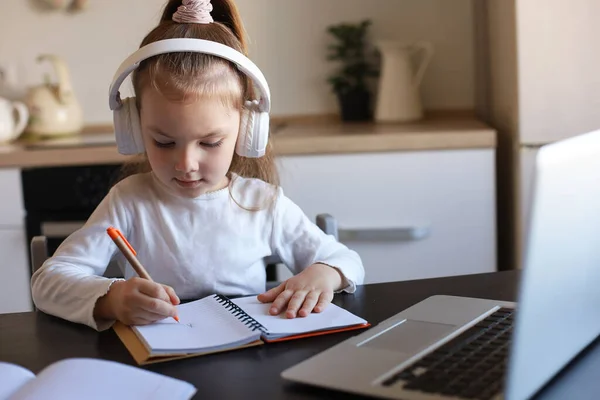 家でオンライン学習ノートブックで机の書き込みに座っているヘッドフォンの女の子 小さな子供の手書き隔離に宿題を準備し ウェブクラスやレッスンを屋内で持っている — ストック写真