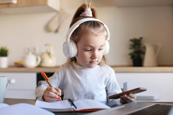 ヘッドフォンのスマート女の子は スマートフォン上で宿題をオンラインクラスを行う インターネット上のイヤホン研究の小さな子供 隔離中にWeb会議やレッスンを持っています — ストック写真