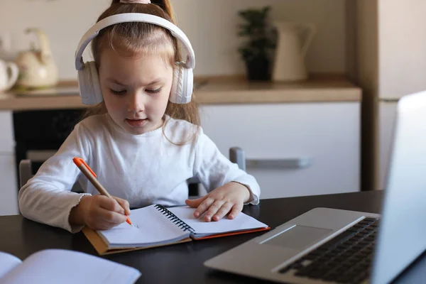 家でオンライン学習ノートブックで机の書き込みに座っているヘッドフォンの女の子 小さな子供の手書き隔離に宿題を準備し ウェブクラスやレッスンを屋内で持っている — ストック写真