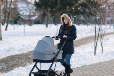 Stoller küçük bebeği ile parkta yürüyüş güzel anne. Kadın mavi jaket hood ve kot pantolon giymiş. Sıcak boots
