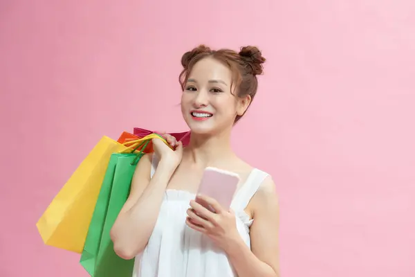 スマートフォンを使いピンクに隔離されたショッピングバッグを持っている幸せな女性 — ストック写真