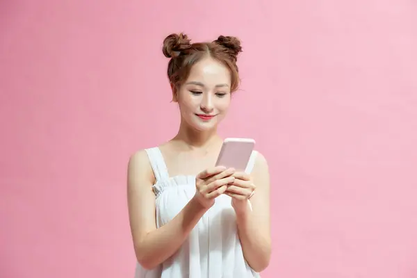 美丽的亚洲女孩使用手机聊天和微笑 站在粉红的背景下 — 图库照片