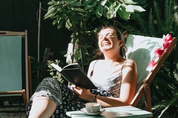 Woman laughing while reading a book while reclined on a chair during a sunny day, copy space, relax and hobby concepts, social network