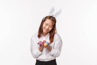 Paskalya tatili, kutlama konsepti. Tavşan kulaklı şirin ve aptal genç Asyalı kız portresi, boyalı yumurtalar ve gülümseyen mütevazı, beyaz arka plan